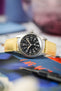 A Hamilton Khaki Field Mechanical watch, featuring a black dial with white Arabic numerals and a 24-hour scale, is fitted with a vibrant yellow Hirsch Duke leather watch strap that has a crocodile-style texture. The watch is positioned on a deep blue book or magazine, with a black pen in the foreground and blurred colored elements in the background.