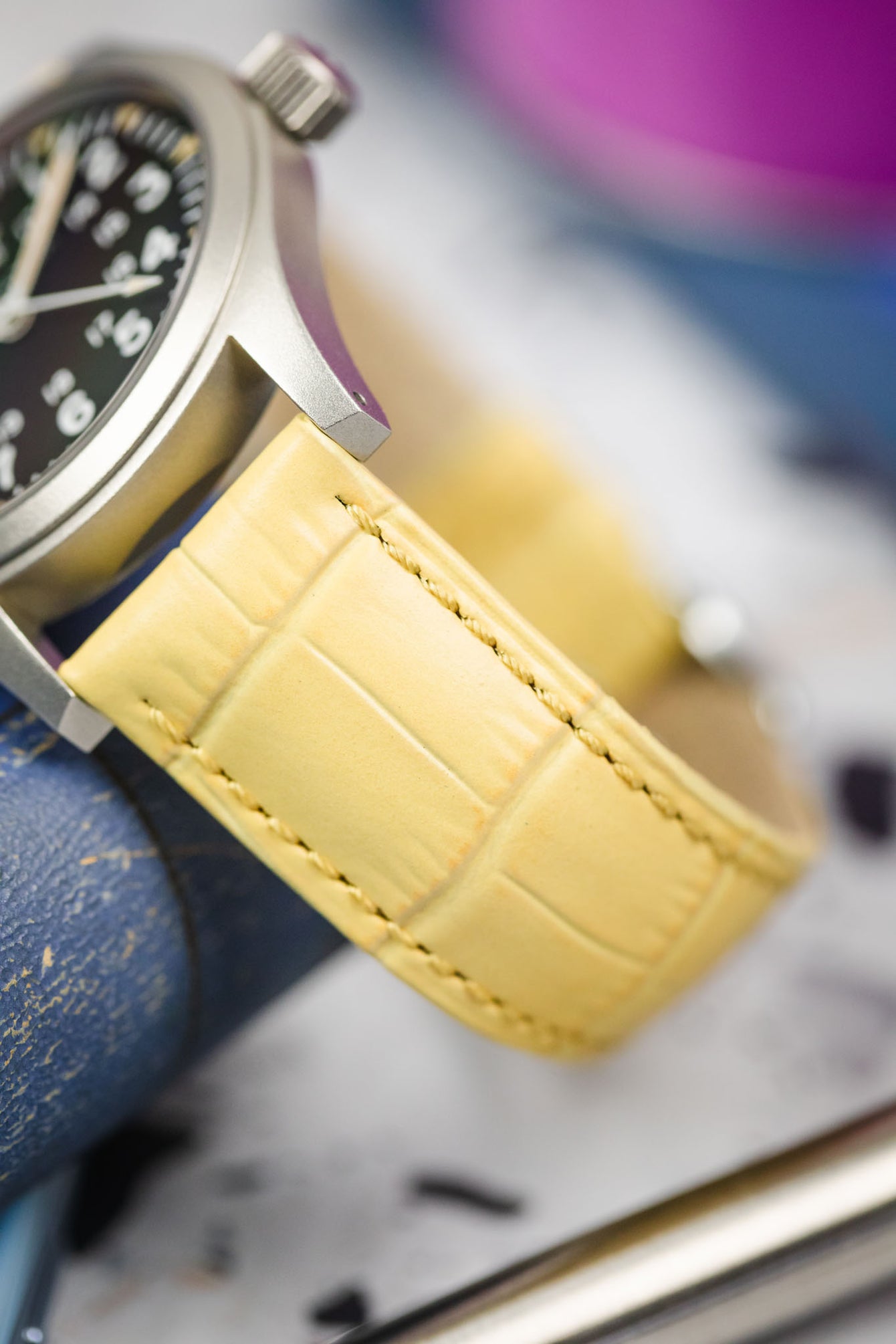 A close-up, eye-level shot of a silver-toned Hamilton Khaki Field Mechanical watch, featuring a black dial with white Arabic numerals and a yellow Hirsch Duke leather watch strap with a textured crocodile-style pattern. The watch is positioned on a blurry, textured surface, with hints of blue and purple in the background.