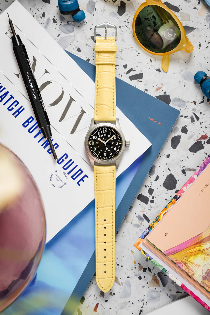 A Hamilton Khaki Field Mechanical watch, featuring a silver-toned case and a black dial with white Arabic numerals, is fitted with a vibrant yellow Hirsch Duke leather strap with crocodile-style embossing. The watch is positioned diagonally on a light grey terrazzo-patterned surface. Surrounding it are a black "WATCH OBSESSION" screwdriver, yellow-tinted sunglasses, blue wireless earbuds, and several publications, including a "WATCH BUYING GUIDE" magazine.