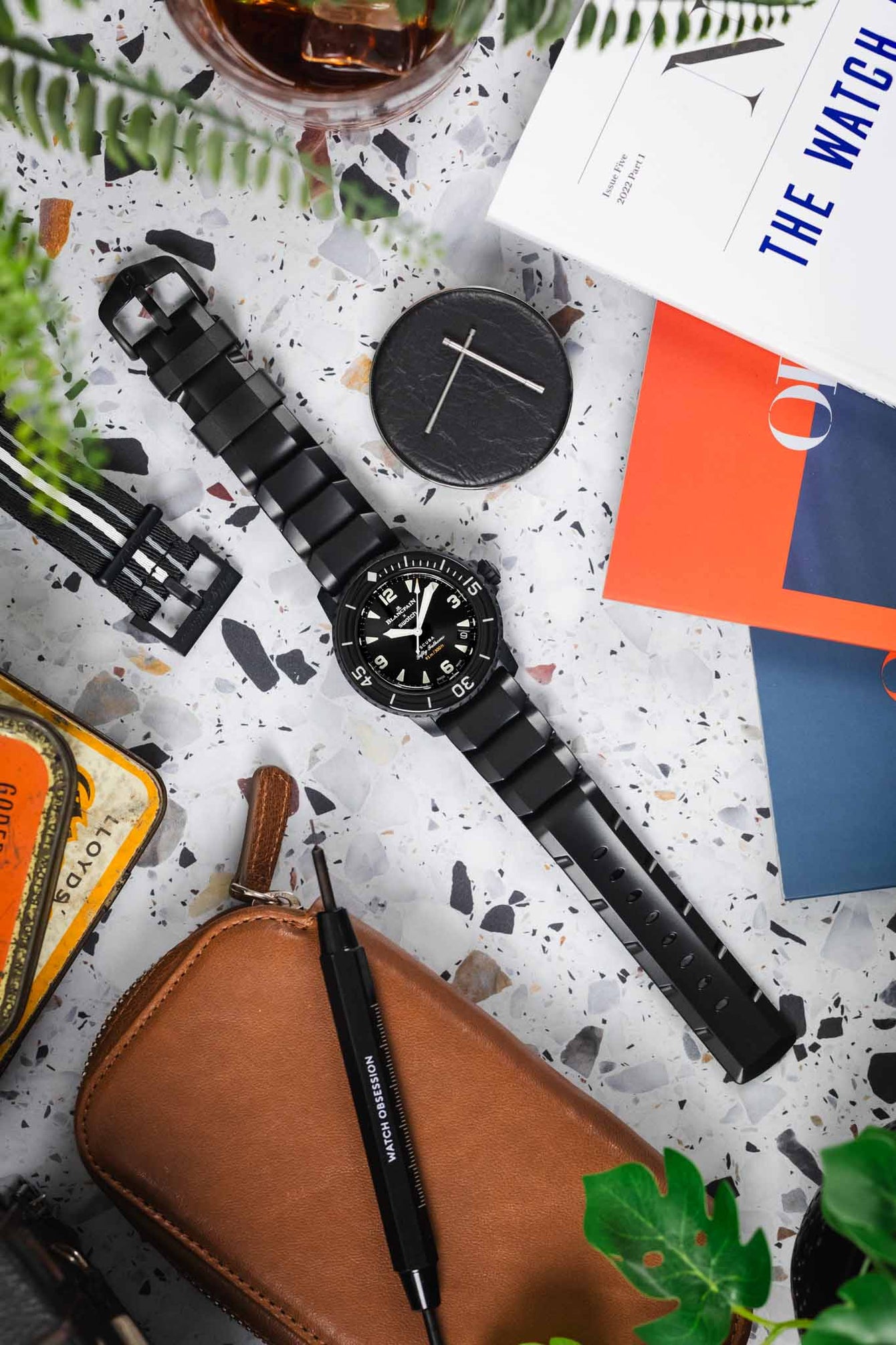 A lay flat photography of a Swatch X Blancpain Fifty Fathoms fitted to a Hirsch Extreme Rubber Diving strap in Black. It is surrounded by watch magazines, tools and a leather watch case against a Terrazzo background.