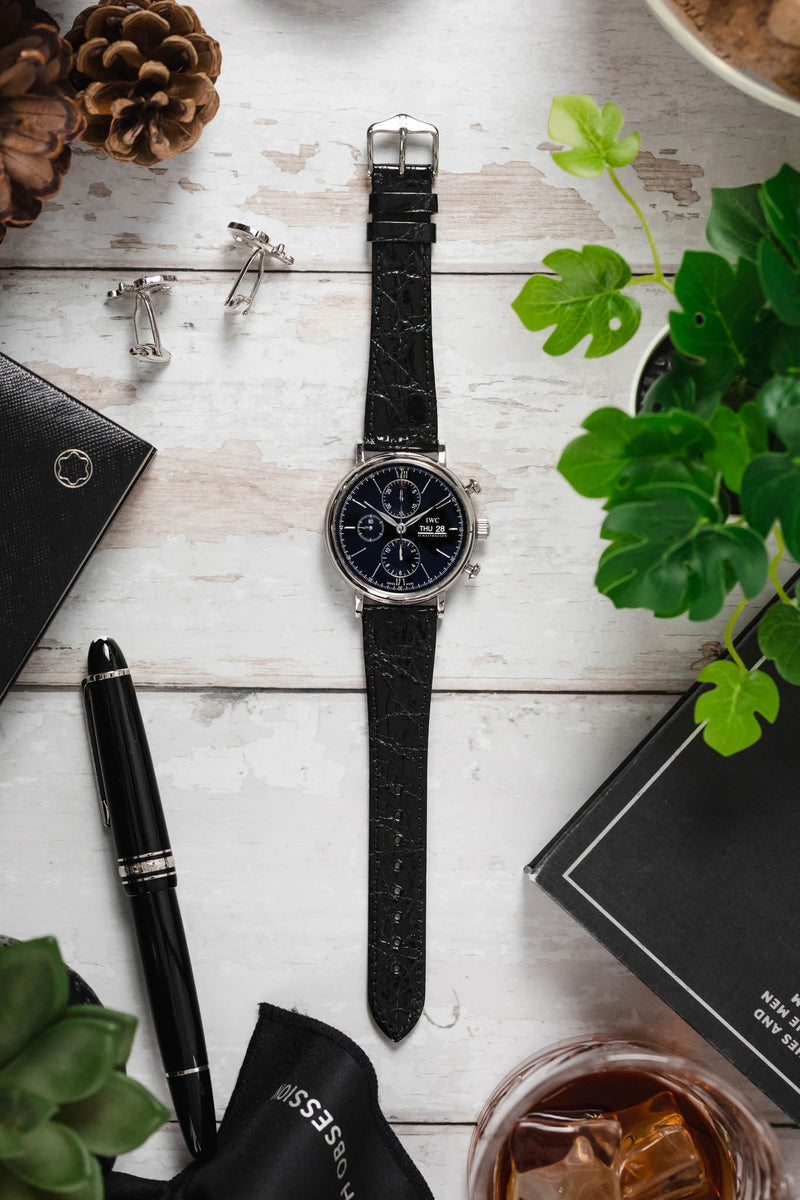 Black crocodile-embossed leather watch strap fitted to an IWC Portofino chronograph watch, displayed with a fountain pen, cufflinks, books, plants, and a whisky glass on a wooden surface.