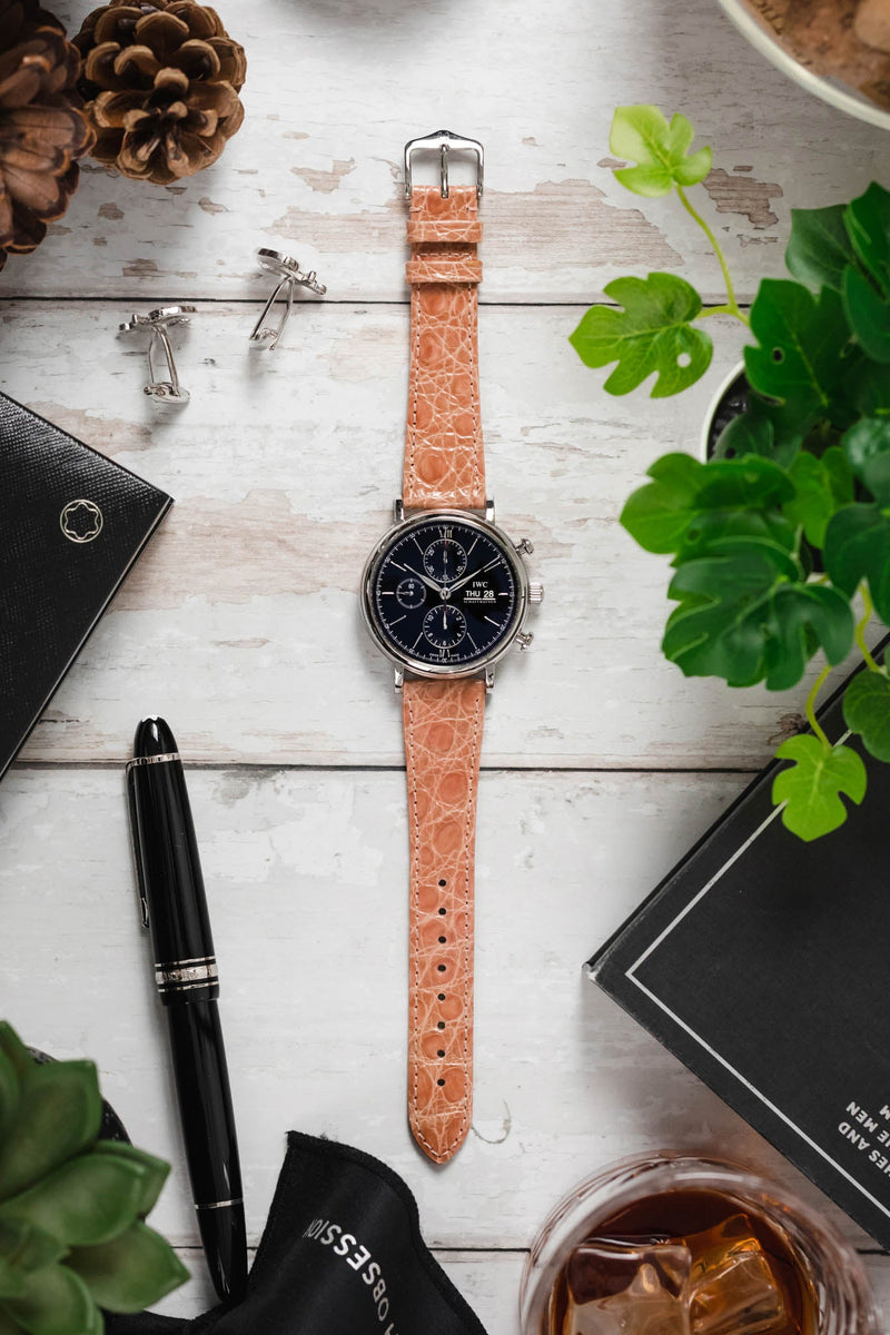 Flat lay of an IWC chronograph watch on a pink crocodile-embossed leather strap, surrounded by a pen, cufflinks, pinecones, greenery, and a glass of amber drink on a rustic white wooden surface.
