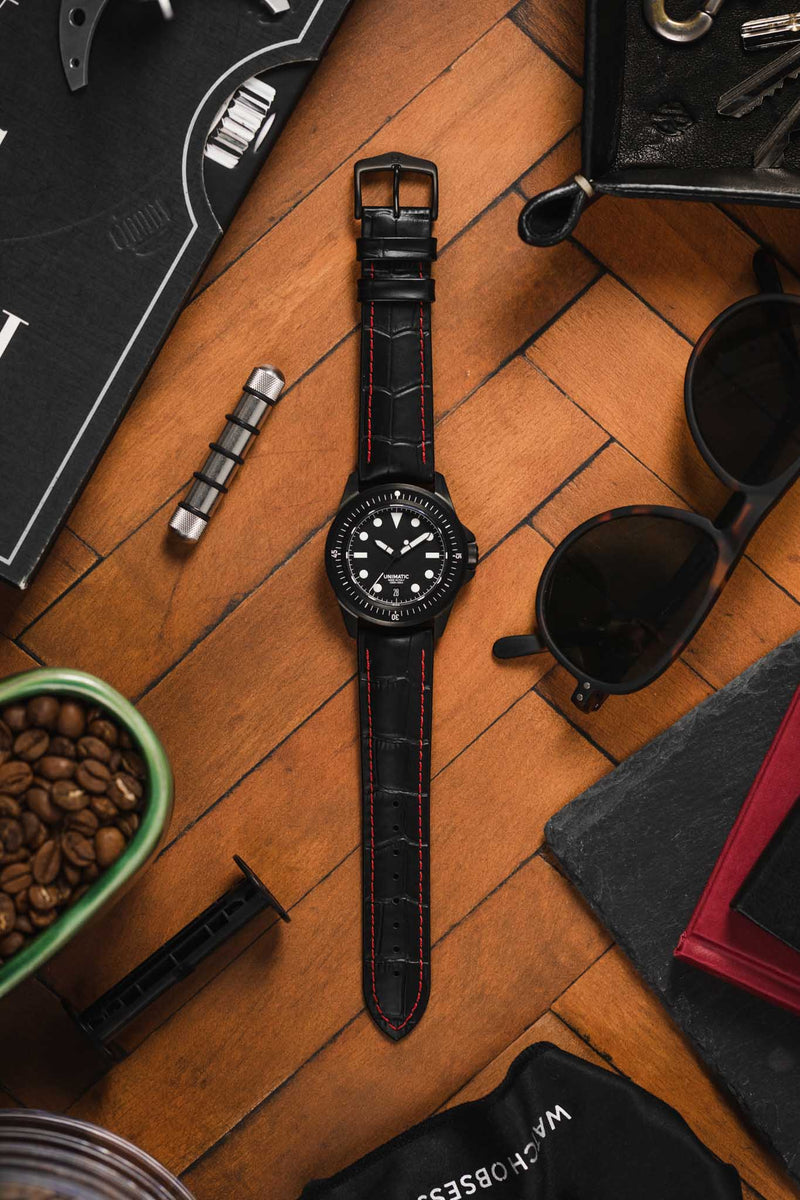 Flat lay of a black dive-style wristwatch with a black dial and white markers, fitted on a black crocodile-embossed leather strap with red contrast stitching, arranged on a wooden surface. Surrounding items include black sunglasses, a bowl of coffee beans, a small metal watch tool, keys in a black leather tray, and books with dark covers, creating a warm, lifestyle-style composition with rich textures and everyday accessories.