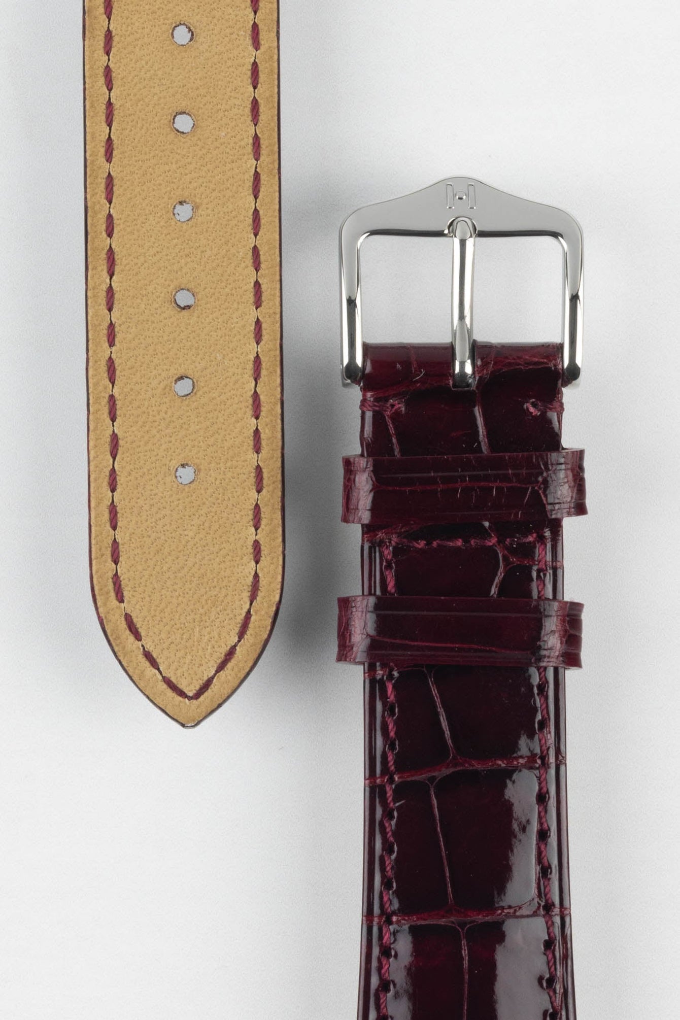 Close-up product image showing both sides of a glossy burgundy crocodile-pattern leather watch strap on a white background, with the front featuring a polished silver-tone pin buckle and two keepers, and the reverse revealing a beige lining with burgundy stitching and evenly spaced adjustment holes.