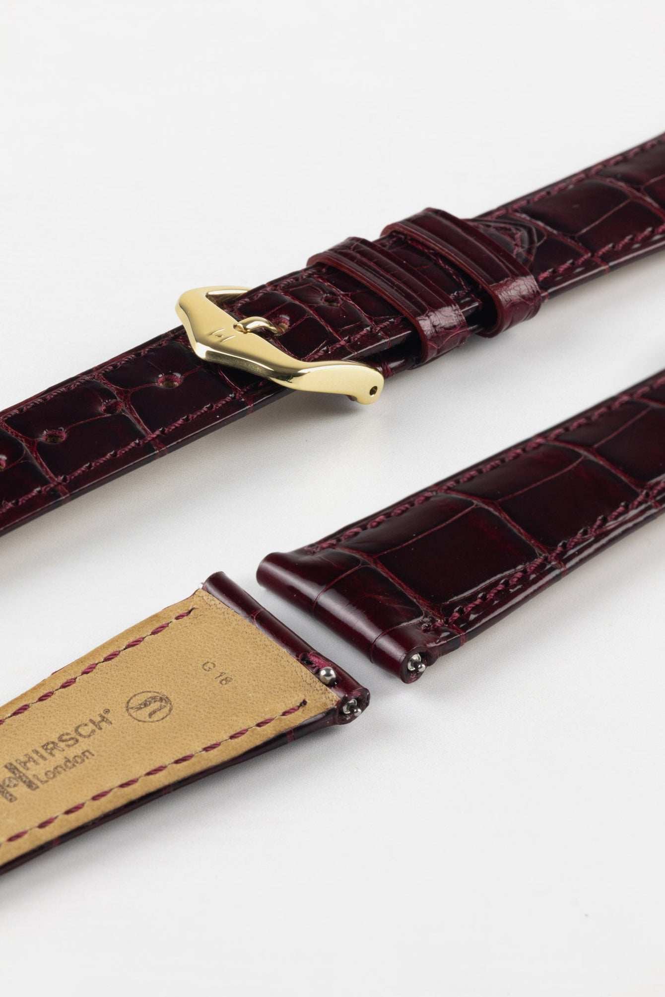 Close-up product image of a glossy burgundy crocodile-pattern leather watch strap with matching stitching, fitted with a polished gold-tone pin buckle, displayed against a white background, with the underside showing beige lining and “HIRSCH London” branding.