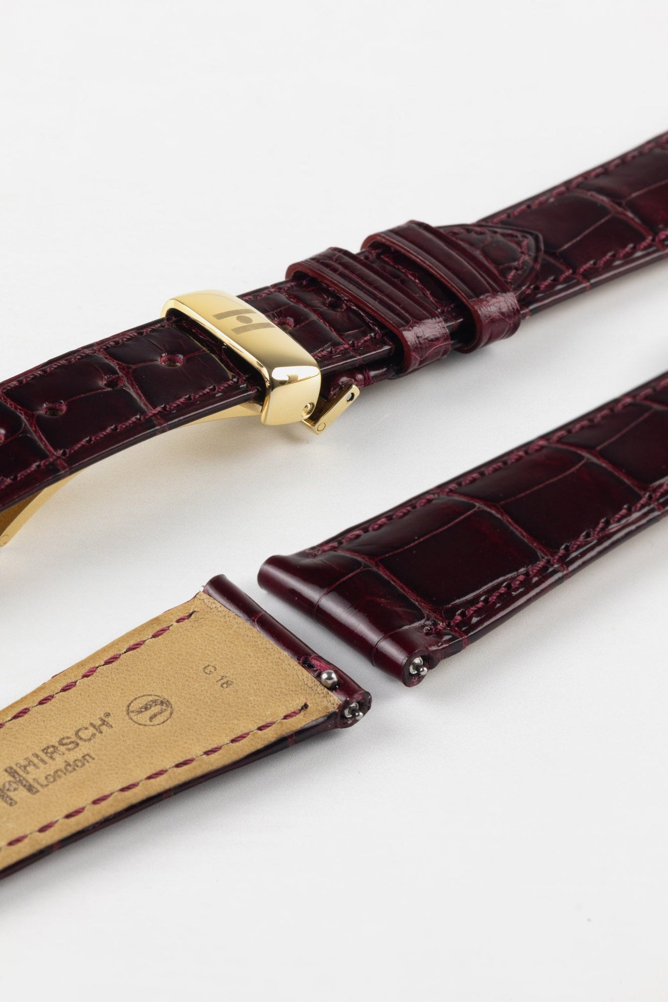 Close-up product image of a glossy burgundy crocodile-pattern leather watch strap with visible stitching, fitted with a polished gold-toned deployant clasp, shown against a white background, with the underside revealing a beige lining stamped “HIRSCH London.”