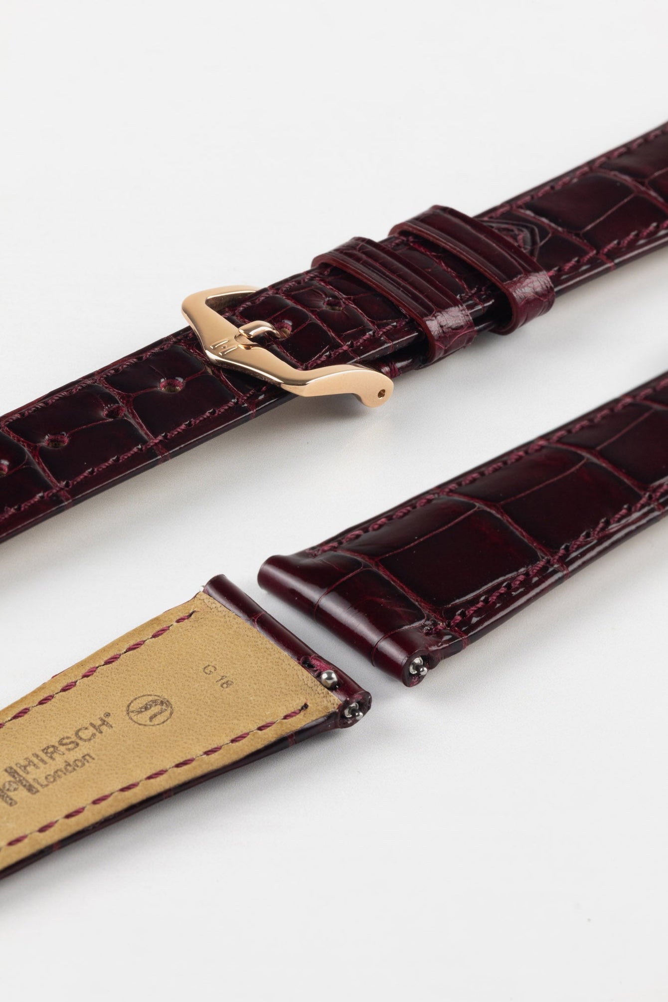 Close-up product image of a glossy burgundy crocodile-pattern leather watch strap with tone-on-tone stitching, featuring a polished rose gold-tone pin buckle, displayed against a white background with the beige underside stamped “HIRSCH London.”