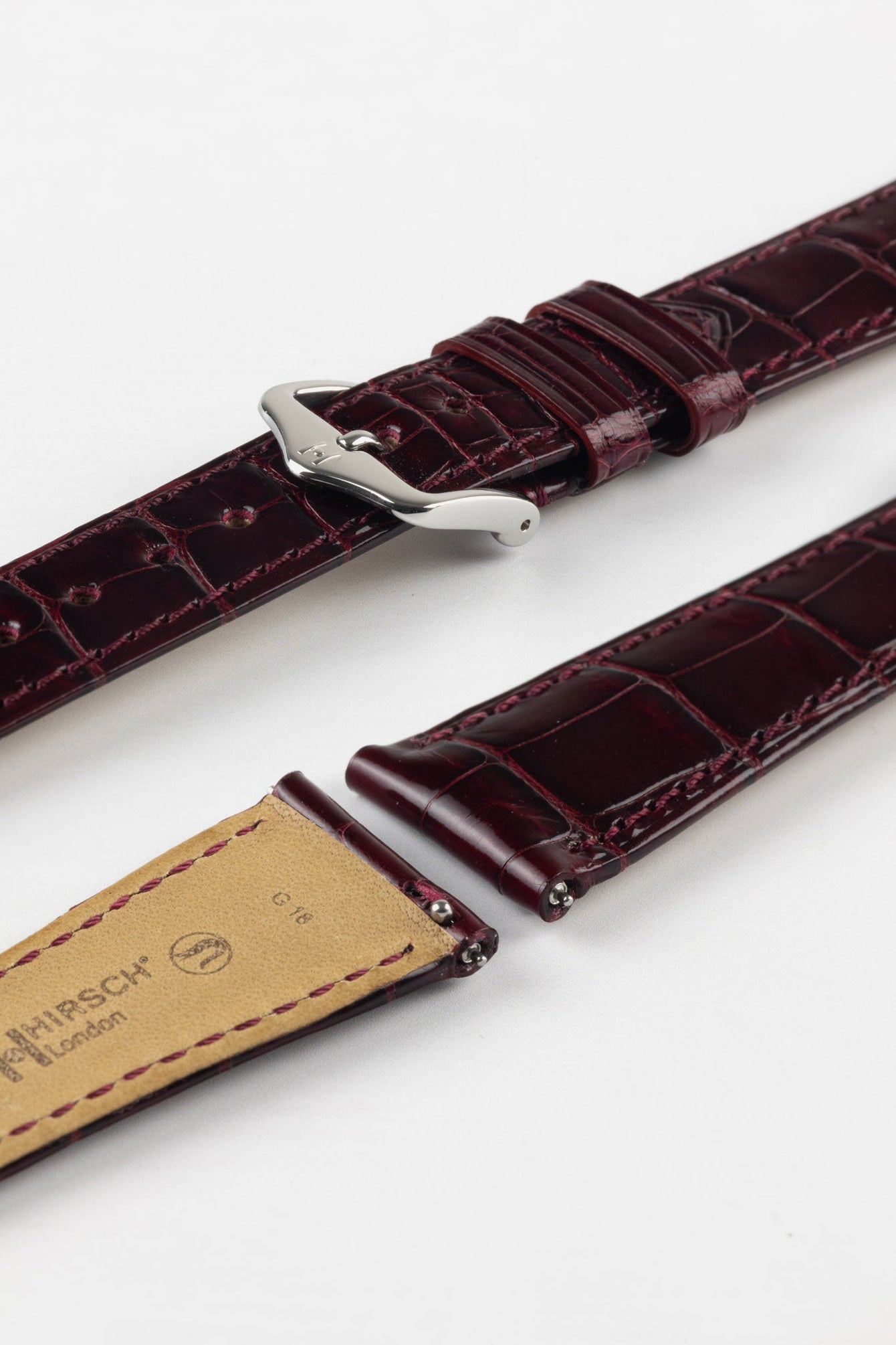 Close-up product image of a glossy burgundy crocodile-pattern leather watch strap with tone-on-tone stitching, featuring a polished gold-tone pin buckle, displayed against a white background with the beige underside stamped “HIRSCH London.”