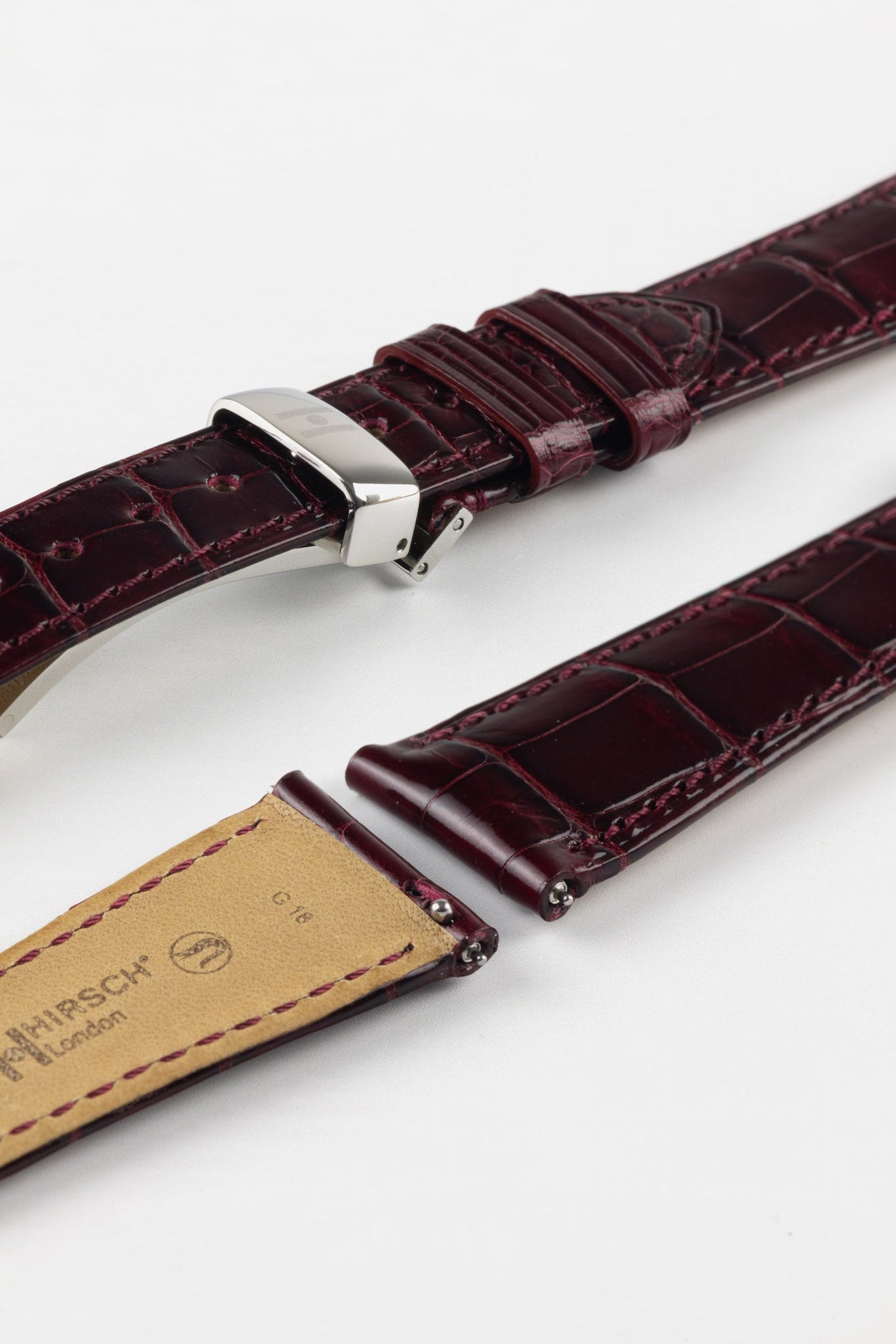 Overhead lifestyle shot of a burgundy crocodile-pattern leather watch strap paired with a white-dial Omega Speedmaster-style chronograph, arranged on a light stone surface alongside a vintage film camera, a pen, magazines, a pinecone, and green plant leaves, with a black cloth and cup partially visible.