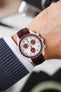 Close-up of a white Omega x Swatch Speedmaster-style chronograph worn on a wrist, featuring a white dial with red sub-dials and a red tachymeter bezel, paired with a glossy burgundy crocodile-pattern leather strap, with a shirt cuff and knit sleeve visible in the foreground.