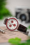 Close-up lifestyle image of a white Omega x Swatch Speedmaster-style chronograph with a white dial and red bezel, paired with a glossy burgundy crocodile-pattern leather strap, resting on a book or magazine, with a blurred camera and green plant in the background.
