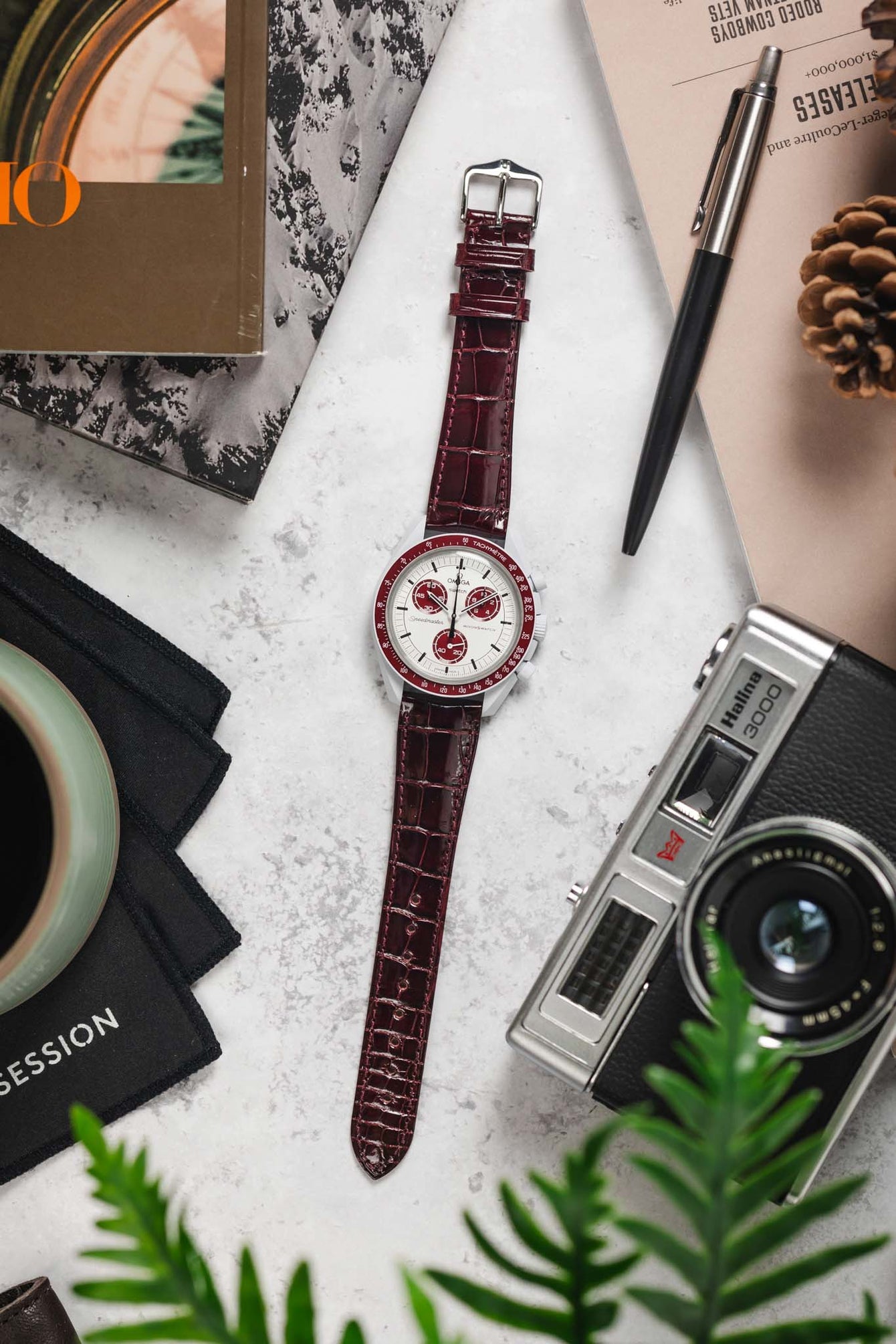 Overhead lifestyle shot of a burgundy crocodile-pattern leather watch strap paired with a white-dial Omega Speedmaster-style chronograph, arranged on a light stone surface alongside a vintage film camera, a pen, magazines, a pinecone, and green plant leaves, with a black cloth and cup partially visible.