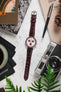 Overhead lifestyle shot of a burgundy crocodile-pattern leather watch strap paired with a white-dial Omega Speedmaster-style chronograph, arranged on a light stone surface alongside a vintage film camera, a pen, magazines, a pinecone, and green plant leaves, with a black cloth and cup partially visible.