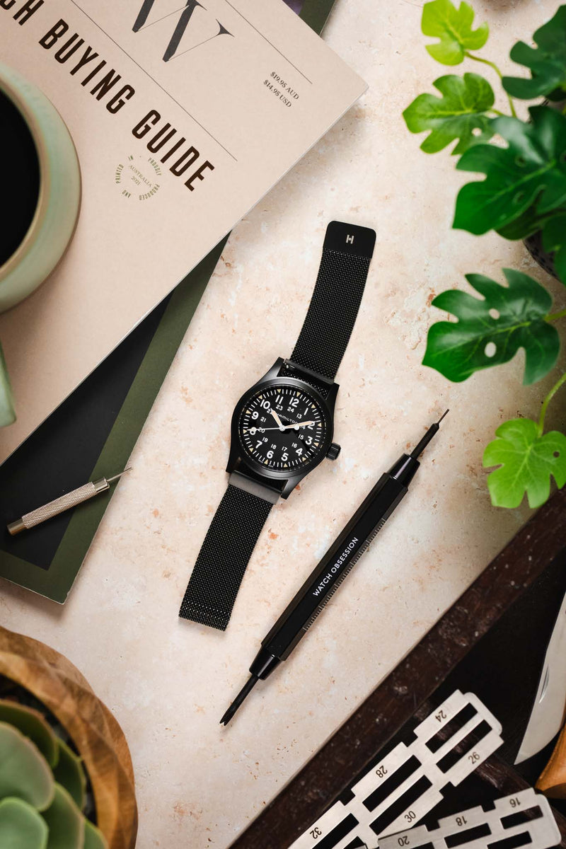 Flat lay of a black watch with a black mesh strap on a light marble surface, surrounded by a watch tool, a “Watch Buying Guide” booklet, and green plant leaves.