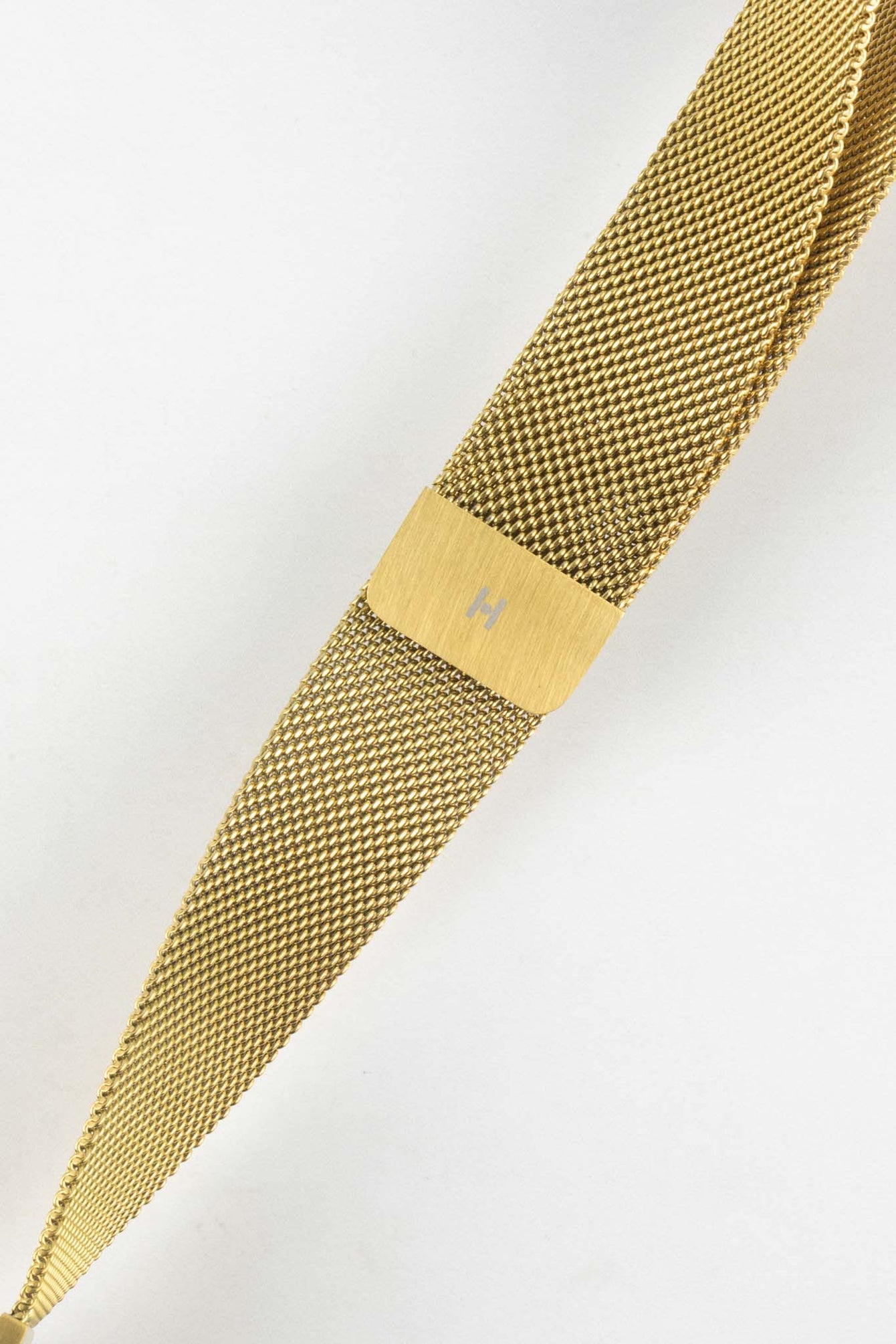 Close-up product image of a gold mesh watch strap against a white background, focusing on the brushed gold sliding clasp with engraved “H” logo, highlighting the fine woven texture and tapered design.