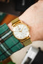 Close-up of a gold-tone Orient dress watch with a white dial and date display worn on a wrist, paired with a gold mesh bracelet, with a green plaid shirt cuff visible and a softly blurred indoor background.