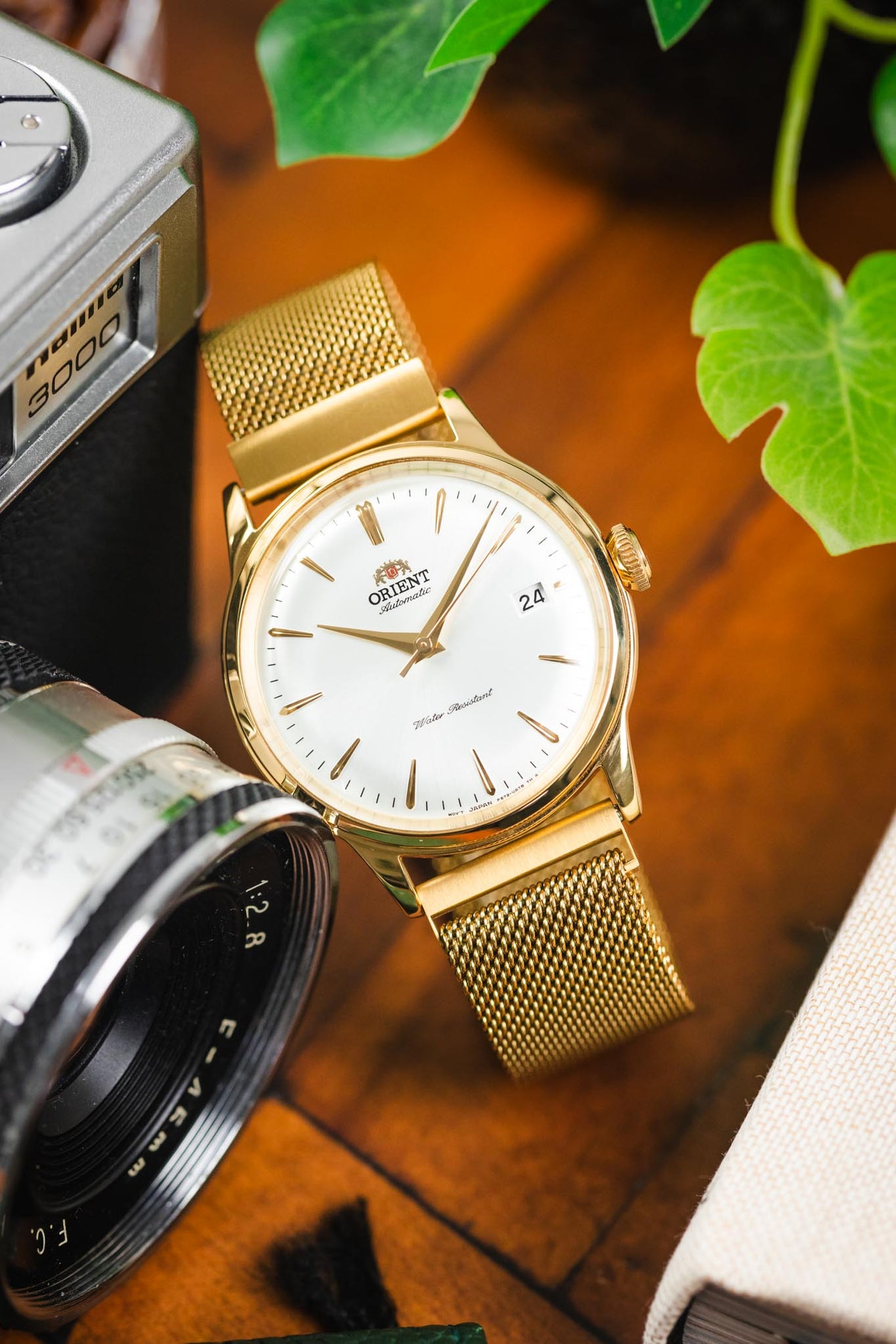 Close-up of a gold-tone Orient dress watch with a white dial and date display, paired with a gold mesh bracelet, resting on a wooden surface beside a vintage camera lens, a small camera, and green plant leaves.