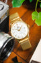 Close-up of a gold-tone Orient dress watch with a white dial and date display, paired with a gold mesh bracelet, resting on a wooden surface beside a vintage camera lens, a small camera, and green plant leaves.