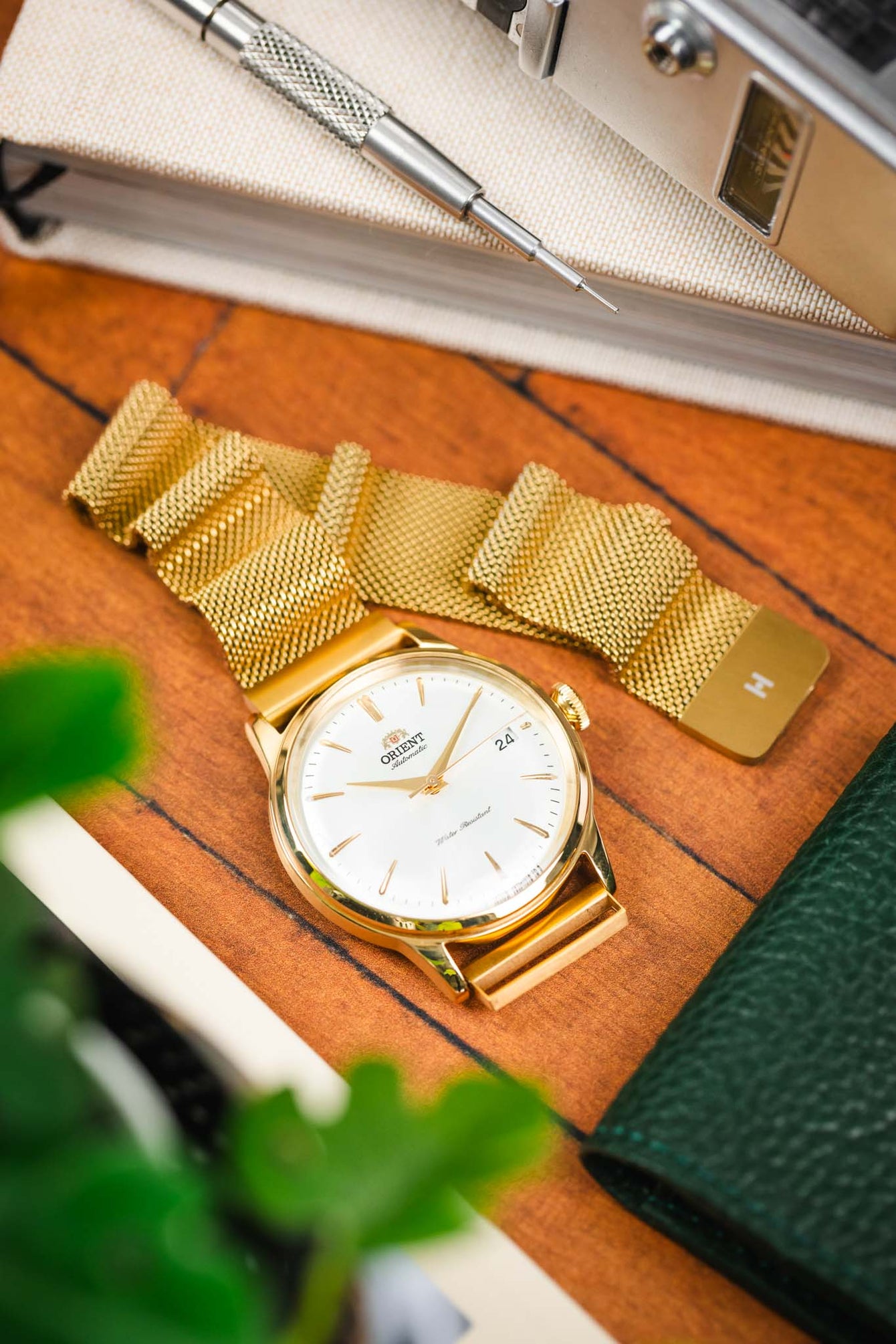 Close-up lifestyle shot of a gold-tone Orient dress watch with a white dial and date display, paired with a gold mesh bracelet, resting on a wooden surface beside a watch tool, a vintage camera, a green leather wallet, and blurred green plant leaves.