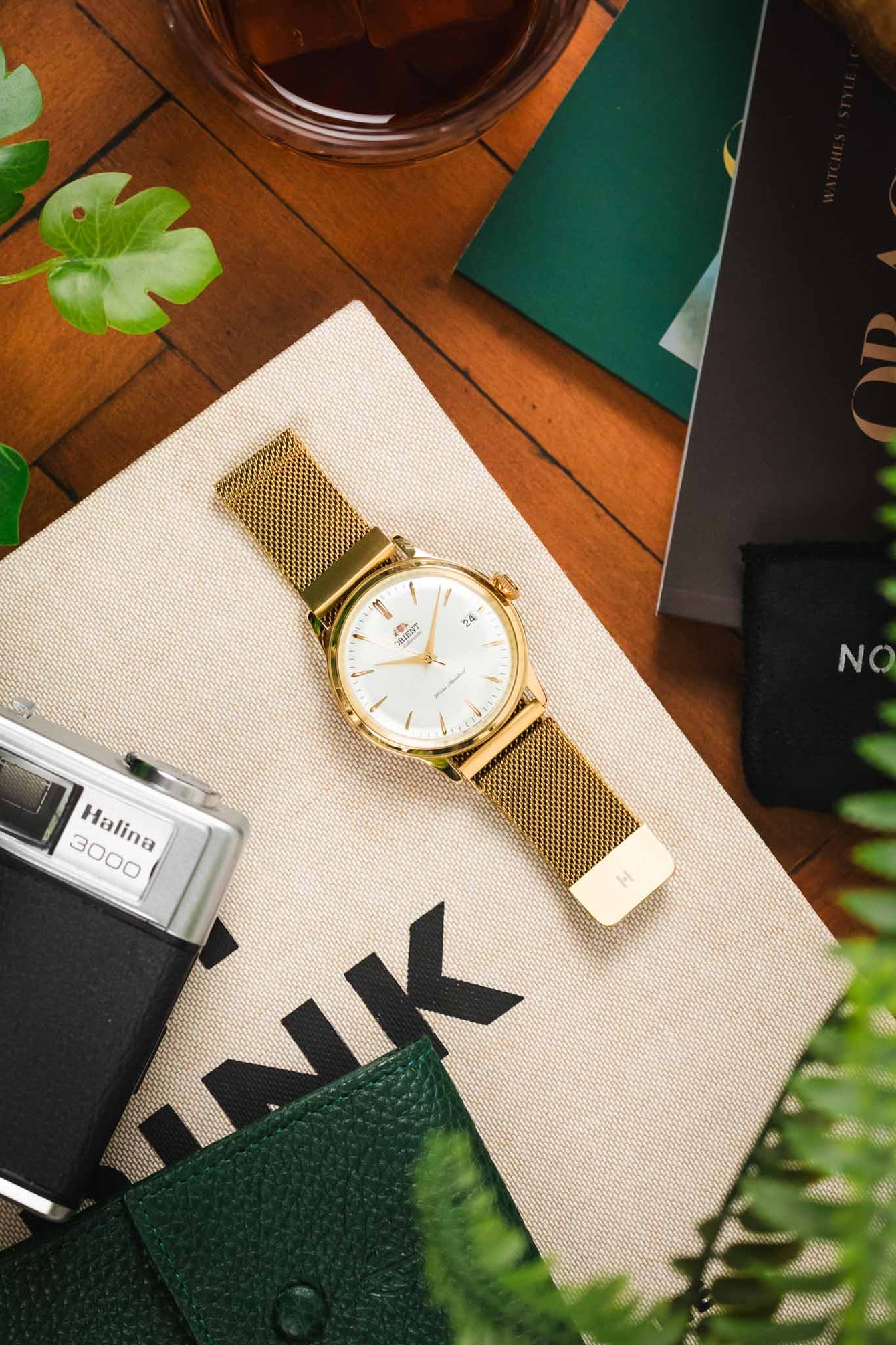 Overhead lifestyle shot of a gold-tone dress watch with a white dial and matching gold mesh bracelet, placed on a canvas tote bag on a wooden surface, surrounded by everyday items including a vintage camera, green leather wallet, magazines, a glass drink, and green plant leaves.