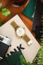 Overhead lifestyle shot of a gold-tone dress watch with a white dial and matching gold mesh bracelet, placed on a canvas tote bag on a wooden surface, surrounded by everyday items including a vintage camera, green leather wallet, magazines, a glass drink, and green plant leaves.