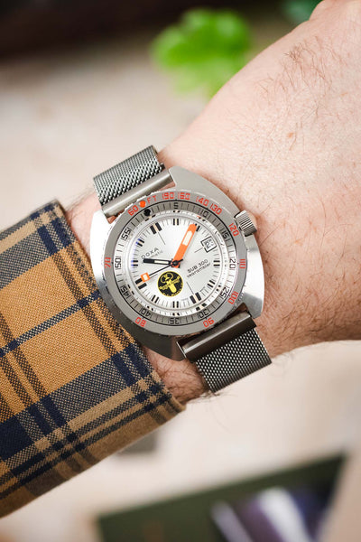 Doxa Sub 300 Searambler with a white dial, orange hand, and mesh bracelet, worn on a wrist with a patterned shirt.