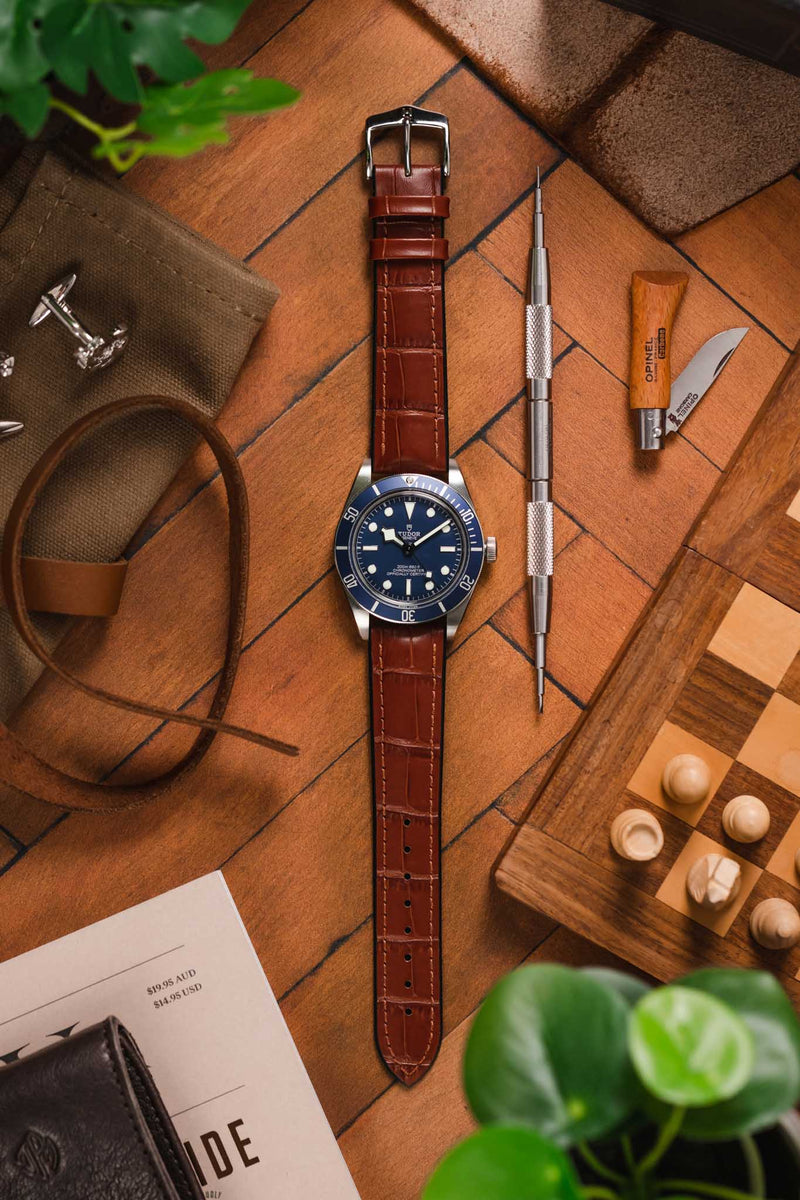 Flat lay of a blue‑dial dive watch on a brown crocodile‑pattern leather strap, surrounded by watch tools, a folding knife, leather accessories, greenery, and a chessboard on a wooden surface.
