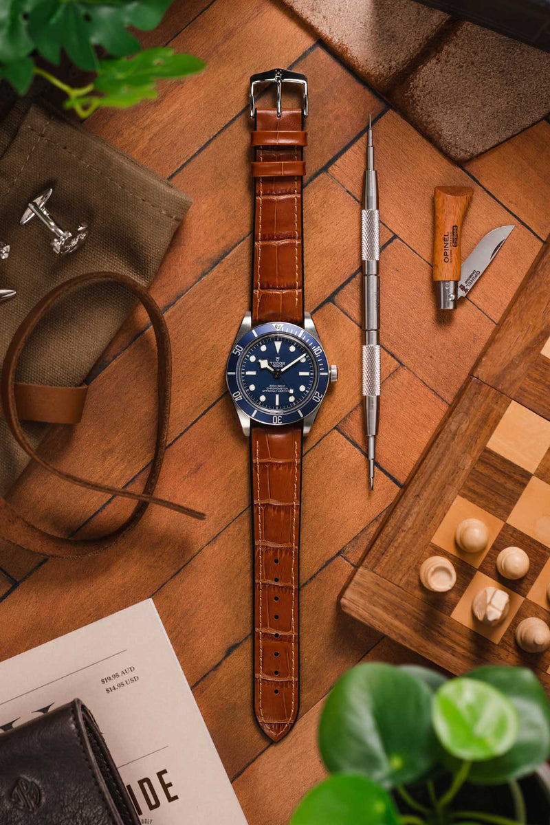 Flat lay of a blue‑dial dive watch on a brown crocodile‑pattern leather strap, arranged on a wooden surface with a spring bar tool, folding knife, leather strap, pouch, chessboard pieces, and greenery.