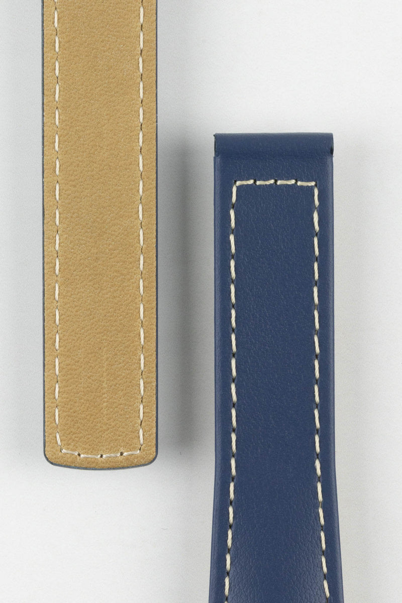 Close-up of a blue calf leather watch strap showing the front and beige underside on a light gray background. The strap features a smooth blue leather finish, white contrast stitching, and padded construction, highlighting the materials and craftsmanship.