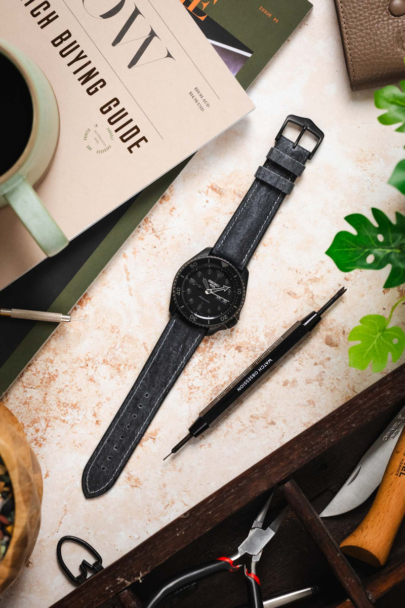 Flat lay of a black dive watch on a black leather strap, surrounded by tools, a coffee cup, greenery, and magazines on a textured surface.