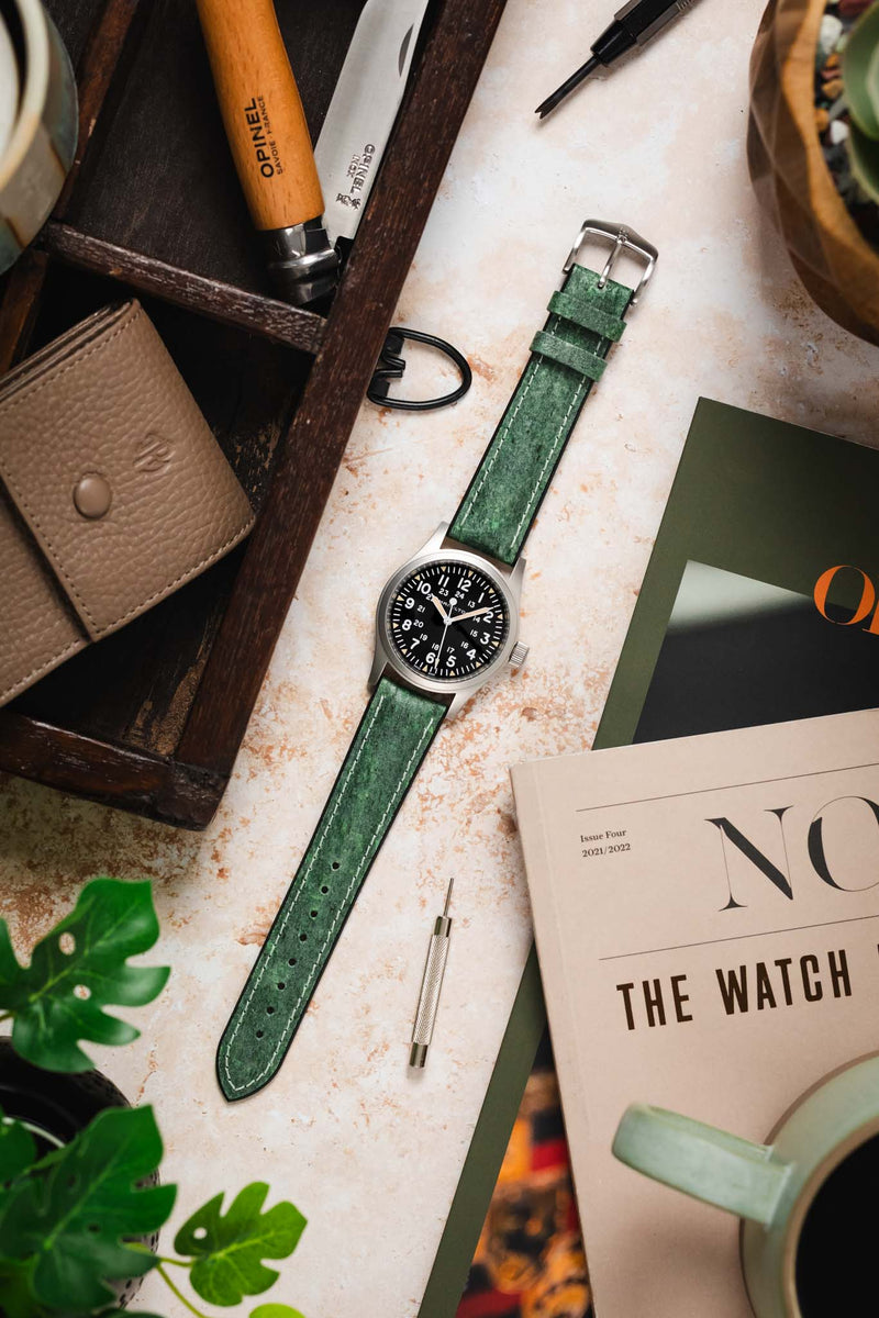 Flat lay of a field watch with a black dial and full Arabic numerals on a green leather strap, arranged on a textured surface. Surrounding items include a wooden tray with tools, a folding knife, a leather pouch, a spring bar tool, magazines, and plant leaves, creating a workshop-style scene.