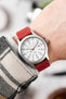 Red leather watch strap fitted to a stainless steel Timex field watch with a white dial, grey Arabic numerals, and orange accent hands, shown on the wrist beneath a cream and black check-pattern jacket. The image highlights the strap’s smooth leather finish, vibrant red colour, comfortable fit, and bold contrast against the brushed steel watch case, demonstrating its casual everyday styling.