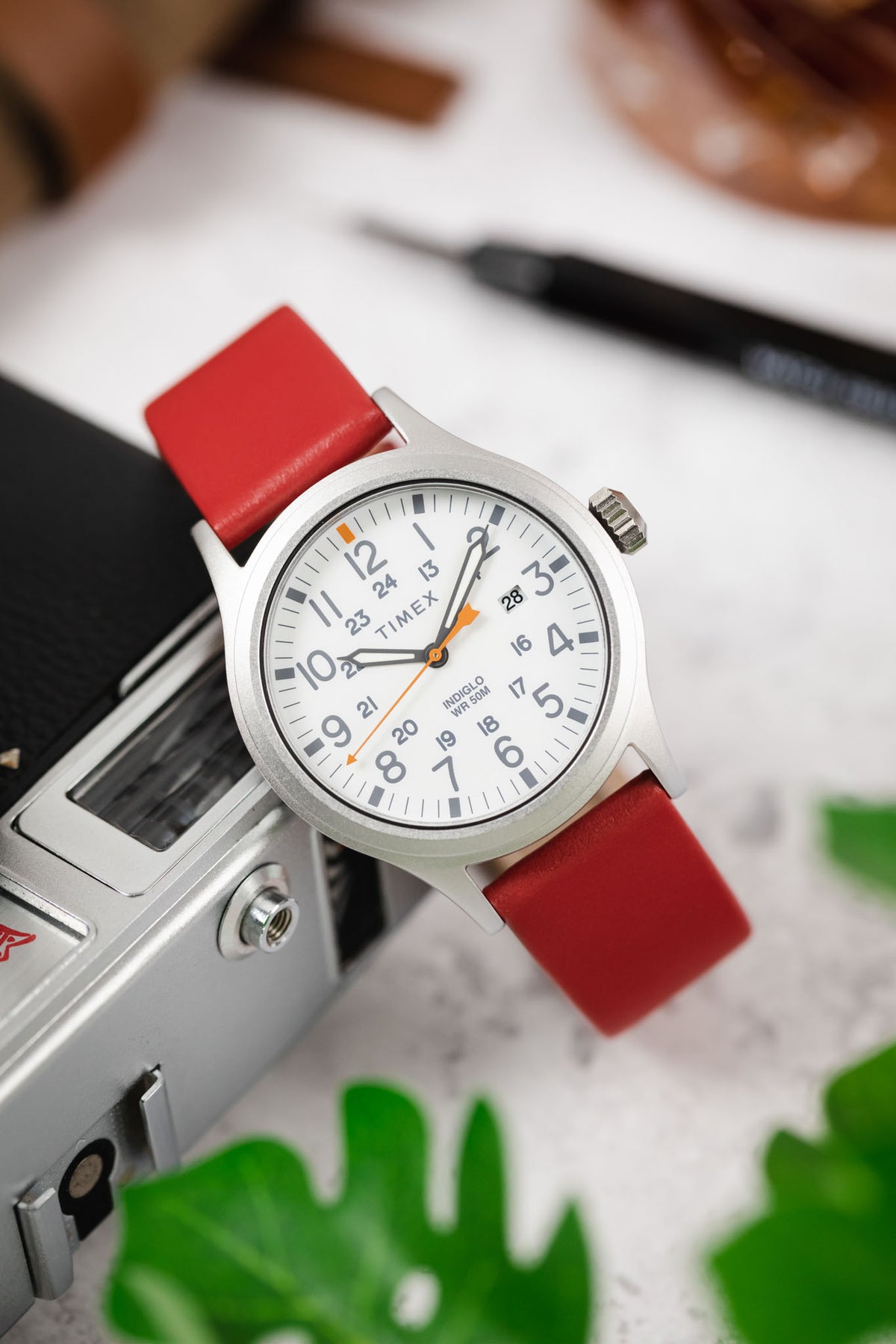 Red leather watch strap fitted to a stainless steel Timex field watch with a white dial, grey Arabic numerals, and orange accent hands, displayed resting on a vintage-style camera. Surrounded by greenery and lifestyle accessories, the image highlights the strap’s smooth leather finish, vibrant red colour, and casual everyday styling in a photography-inspired setting.