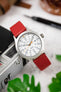 Red leather watch strap fitted to a stainless steel Timex field watch with a white dial, grey Arabic numerals, and orange accent hands, displayed resting on a vintage-style camera. Surrounded by greenery and lifestyle accessories, the image highlights the strap’s smooth leather finish, vibrant red colour, and casual everyday styling in a photography-inspired setting.