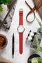 Red leather watch strap fitted to a stainless steel Timex field watch with a white dial and Arabic numeral markers, displayed on a light stone-textured surface alongside a vintage camera, watchmaker’s tool, iced drink, greenery, and a leather notebook. The image highlights the strap’s smooth leather finish, vibrant red colour, and versatile casual styling in a lifestyle-inspired setting.