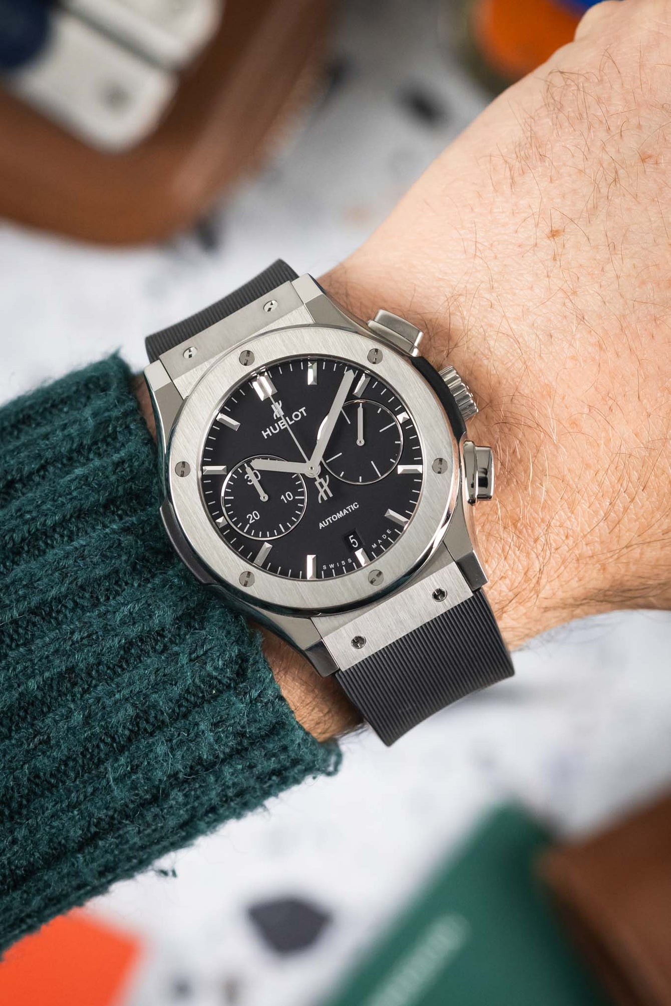 Close-up of a Hublot Classic Fusion chronograph with a black dial and rubber strap, worn on a wrist with a green knit sleeve visible.