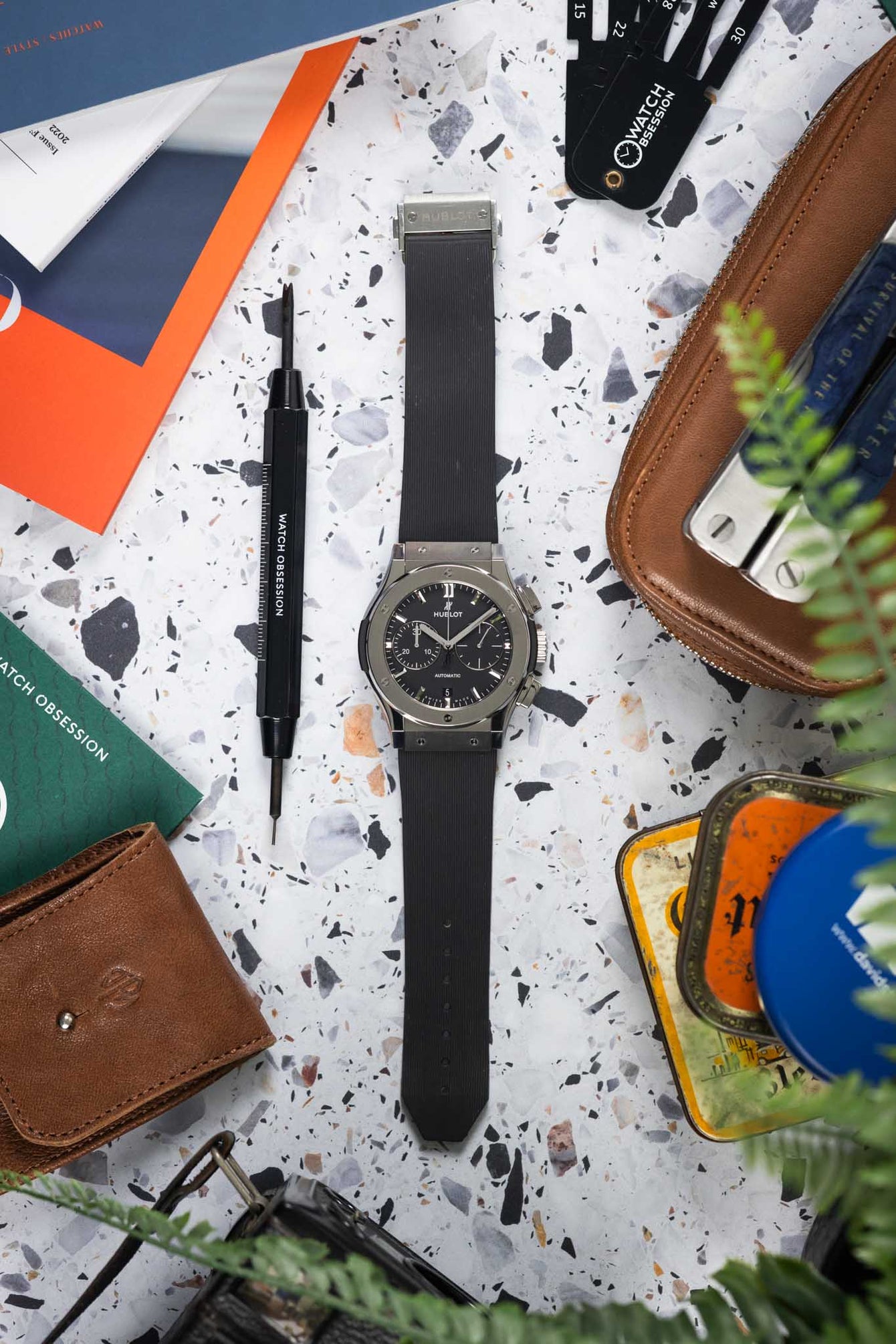 Flat-lay of a Hublot Classic Fusion chronograph on a black rubber strap, arranged on a speckled surface with a watch tool, leather pouch, cards, and greenery.