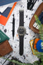 Flat-lay of a Hublot Classic Fusion chronograph on a black rubber strap, arranged on a speckled surface with a watch tool, leather pouch, cards, and greenery.