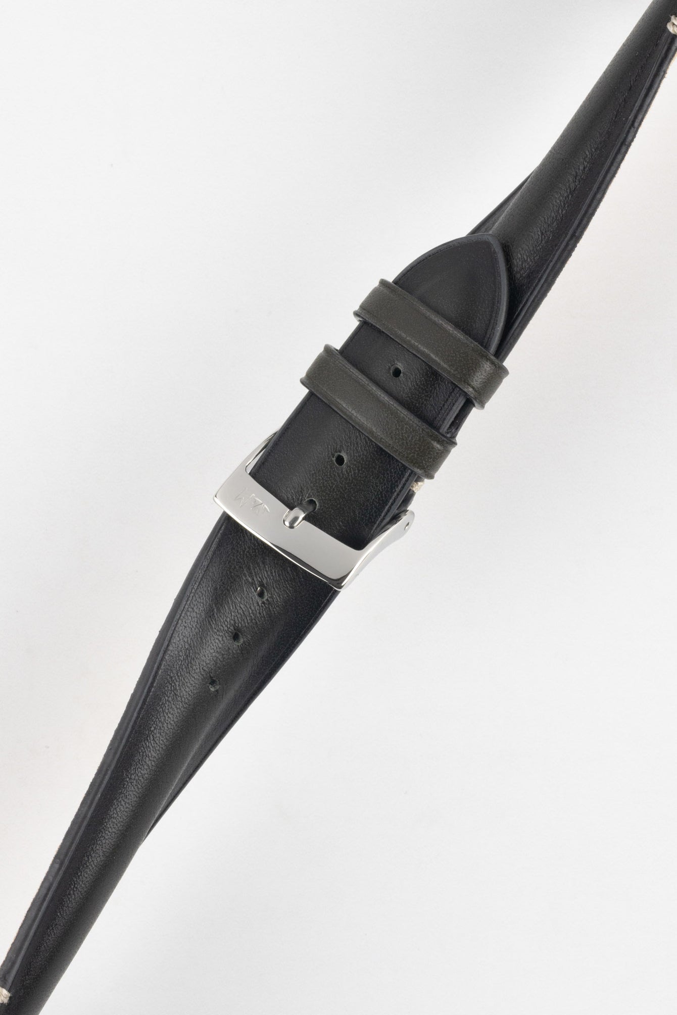 Angled close-up of a black leather watch strap on a light grey background, showing a polished silver tang buckle with an engraved logo, two leather keepers, and a series of adjustment holes along the strap. The leather has a smooth, slightly pebbled texture with subtle creasing and light contrast stitching near the buckle.