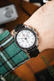 Close-up of an Omega Speedmaster chronograph with a white dial and black bezel, worn on a wrist with a black leather strap and a green plaid sleeve visible.