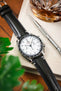 Close-up of an Omega Speedmaster with a white dial and black bezel on a black leather strap, placed beside a whisky glass and watch tool on a fabric surface.