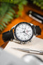 Close-up of an Omega Speedmaster with a white dial and black bezel on a black leather strap, resting on a fabric surface with a blurred background of greenery and desk items.