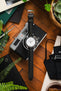 Flat-lay of an Omega chronograph watch on a black leather strap, placed over a camera and surrounded by books, tools, greenery, and a drink on a wooden surface.