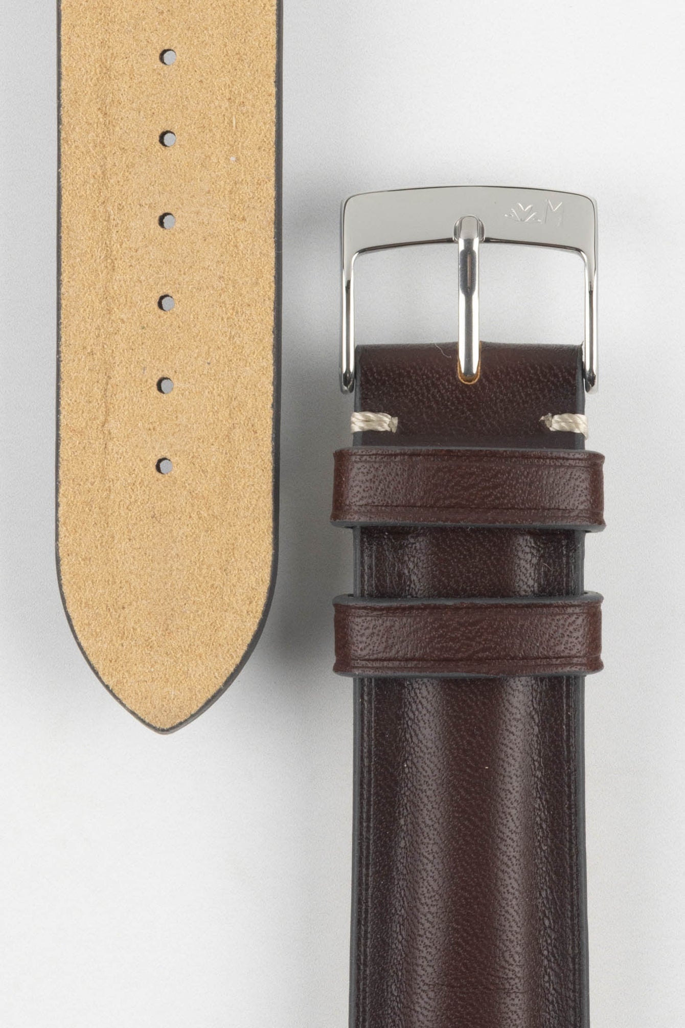 Close-up of a burgundy leather watch strap on a light grey background, showing one section with a polished silver tang buckle featuring an engraved logo and two leather keepers. Beside it, the opposite strap section displays the tan suede underside with a row of evenly spaced adjustment holes. The leather has a smooth, slightly pebbled texture with light contrast stitching near the buckle, highlighting the strap’s construction and finish.