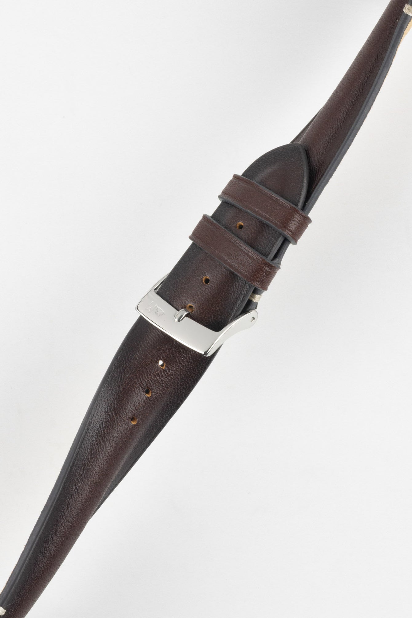 Angled close-up of a burgundy leather watch strap on a light grey background, featuring a polished silver tang buckle with an engraved logo and two leather keepers. The strap shows a smooth, slightly pebbled finish with subtle tonal variation and a row of evenly spaced adjustment holes along its length. The tapered design and visible stitching near the lugs highlight the strap’s refined craftsmanship and premium construction.