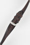 Angled close-up of a burgundy leather watch strap on a light grey background, featuring a polished silver tang buckle with an engraved logo and two leather keepers. The strap shows a smooth, slightly pebbled finish with subtle tonal variation and a row of evenly spaced adjustment holes along its length. The tapered design and visible stitching near the lugs highlight the strap’s refined craftsmanship and premium construction.