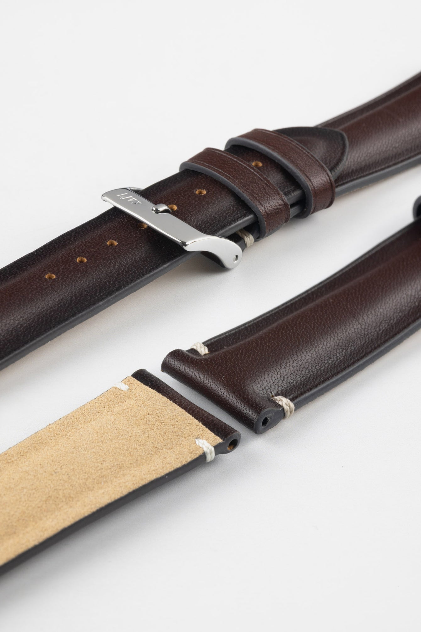 Angled close-up of a burgundy leather watch strap displayed on a light grey background, with the strap separated into multiple sections. One section features a polished silver tang buckle with an engraved logo and two matching leather keepers, while adjacent pieces show the smooth, slightly pebbled burgundy leather exterior and the tan suede underside near the lugs. Evenly spaced adjustment holes and visible stitching highlight the strap’s craftsmanship and premium construction.