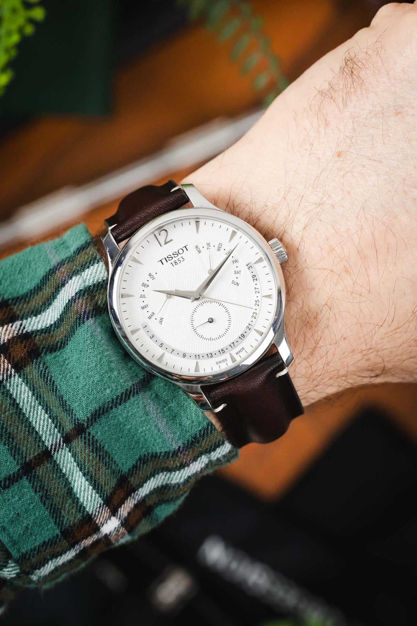 Close-up of a Tissot watch with a white dial and brown leather strap, worn on a wrist with a green plaid sleeve visible.