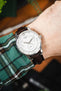 Close-up of a Tissot watch with a white dial and brown leather strap, worn on a wrist with a green plaid sleeve visible.