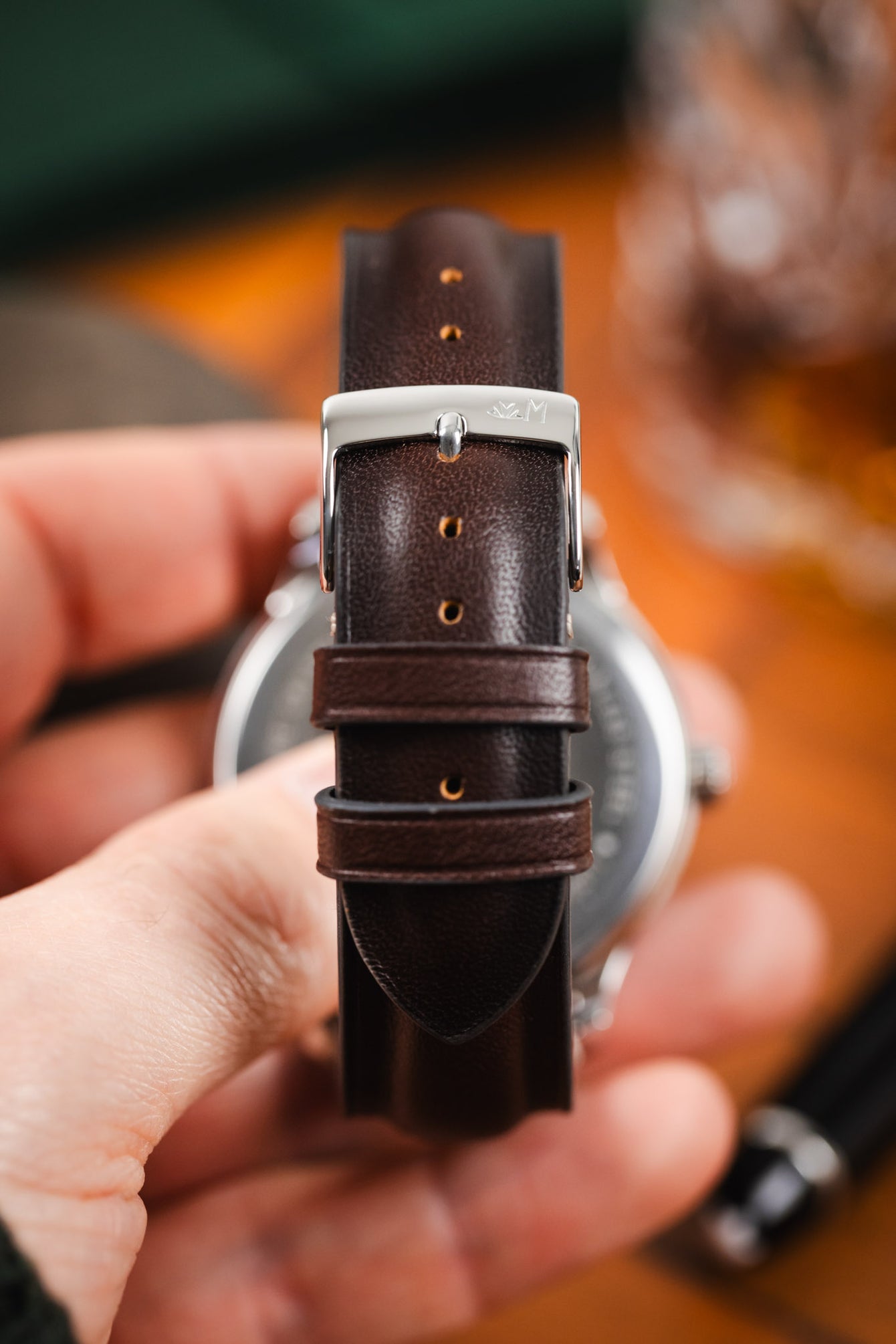 Close-up of a brown leather watch strap with a polished silver buckle, showing adjustment holes and keeper loops while held in hand, with a softly blurred background.